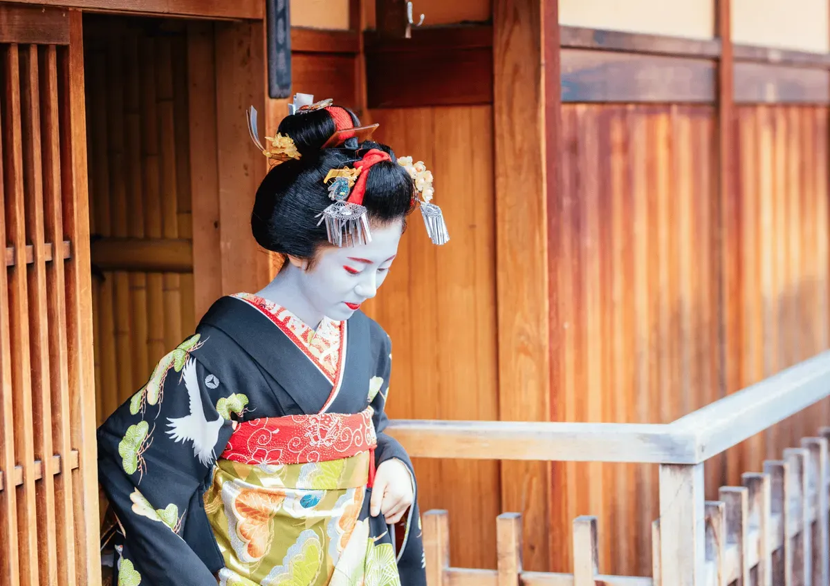 Maiko in a black crane-patterned kimono and silver kanzashi hairpins outside a wooden Gion teahouse in Kyoto