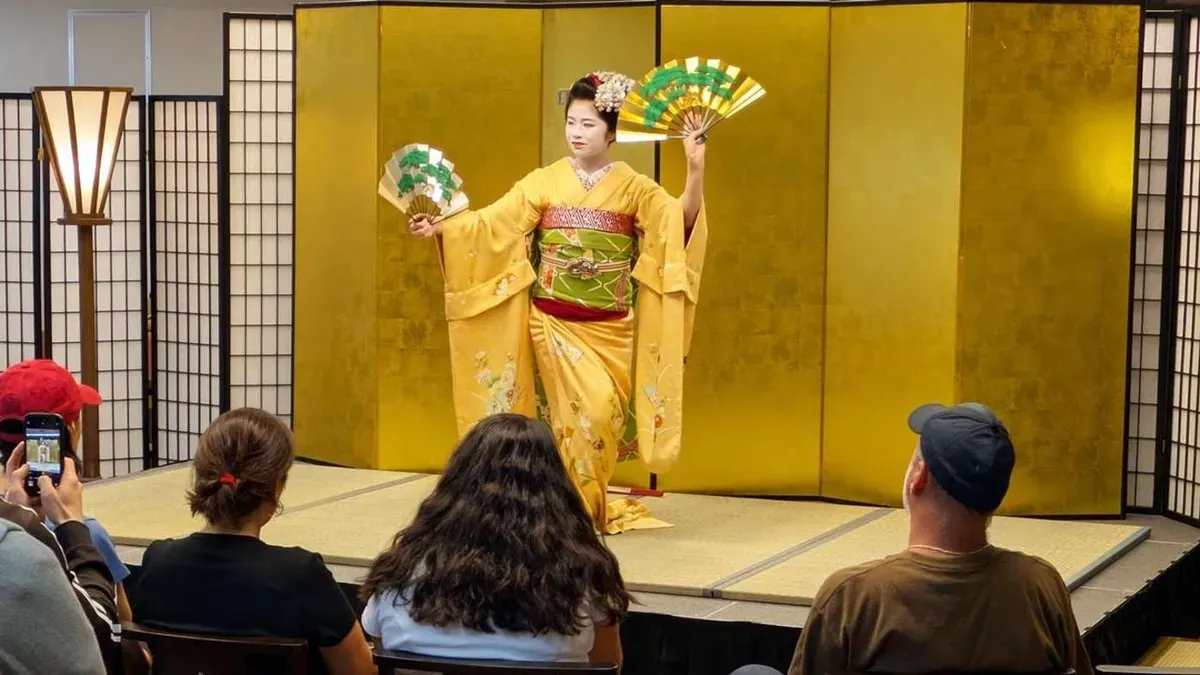 Maiko in a golden kimono performing a fan dance on a tatami stage as guests watch and take photos at a maiko show in Kyoto